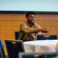 Two men sitting, one talking and gesturing to a room